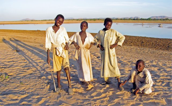 Photo of four young boys standing on a sandy riverbank.